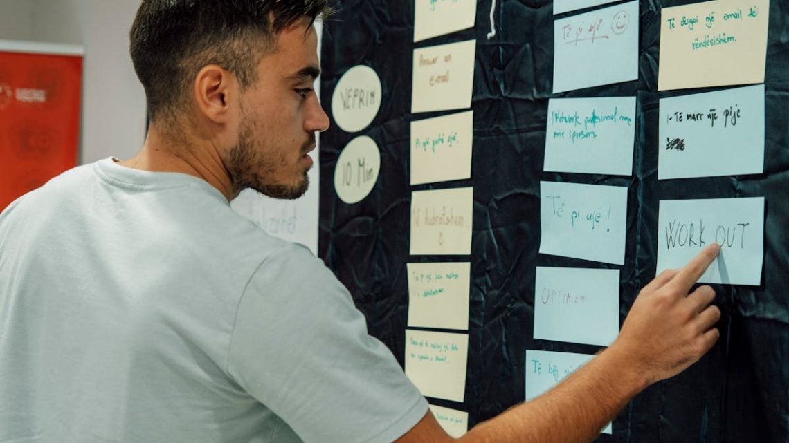 A man in a casual t-shirt organizes notes on a wall, focusing on a 'WORK OUT' sign.